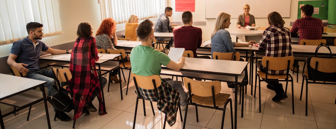 Students sit in pairs at desks in a classroom, facing their instructor.